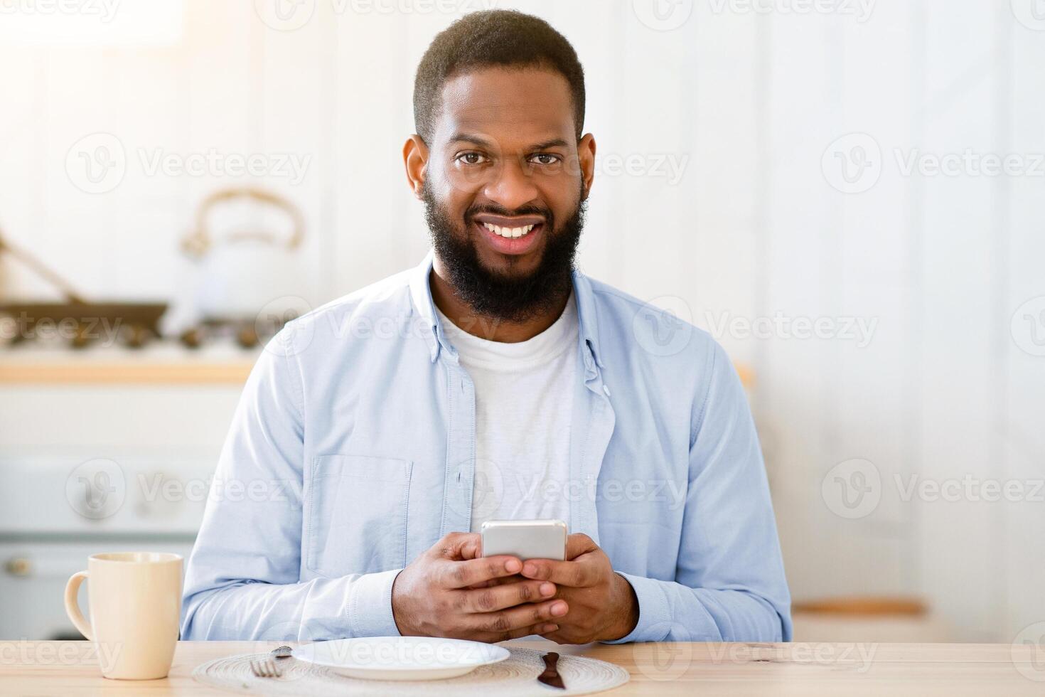 Young African Guy Sitting At Table In Kitchen With Smartphone While His Wife Cooking Lunch On Background, Black Millennial Couple Spending Time Together At Home, Selective Focus With Free Space photo