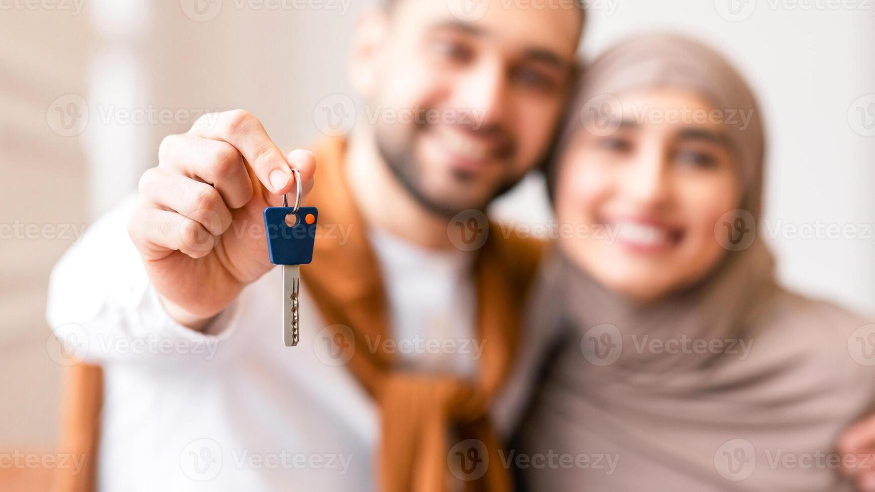 Joyful Muslim Couple Showing New House Key Standing In Their Own Home After Relocation, Focus On Key. Real Estate Ownership And Mortgage Loan, Buying New Flat Concept. Shallow Depth photo