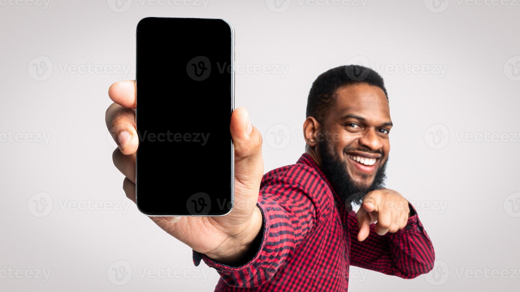 Use This Application. Cheerful Black Guy Showing Phone Empty Screen To Camera Recommending New Mobile App Posing Standing In Studio Over White Background. Smartphone Advertisement. Mockup photo