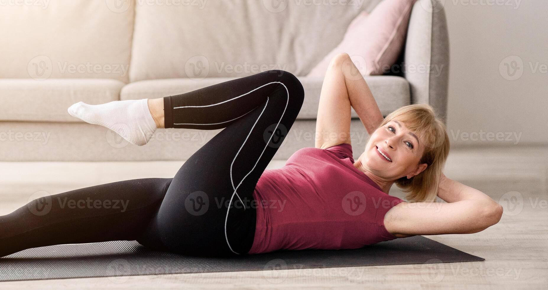 Fit senior woman doing abs exercises on yoga mat indoors, empty space. Full length portrait of sporty mature lady working out at home during coronavirus epidemic. Strength training concept photo