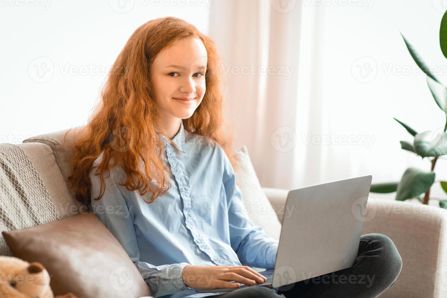 Homeland Generation. Cheerful young ginger hair teenage girl sitting on couch in bedroom, using laptop computer, typing on keyboard, surfing web and looking at camera, resting and relaxing photo