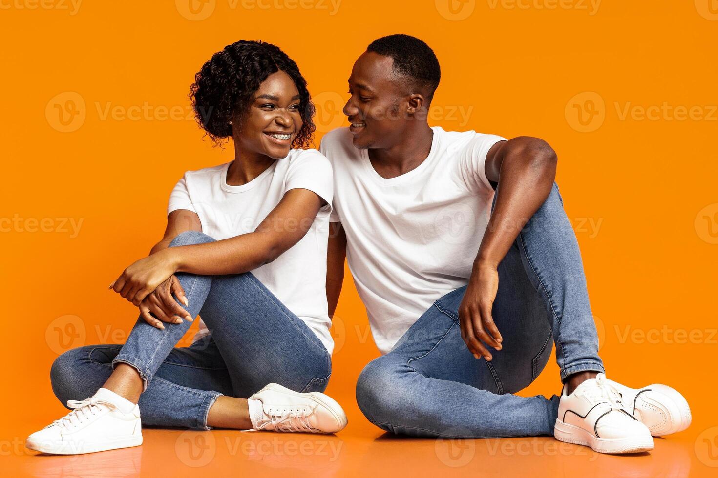 Happy african american lovers sitting on floor over orange studio background, looking at each other, copy space. Loving young black man and woman spending time together, bonding photo