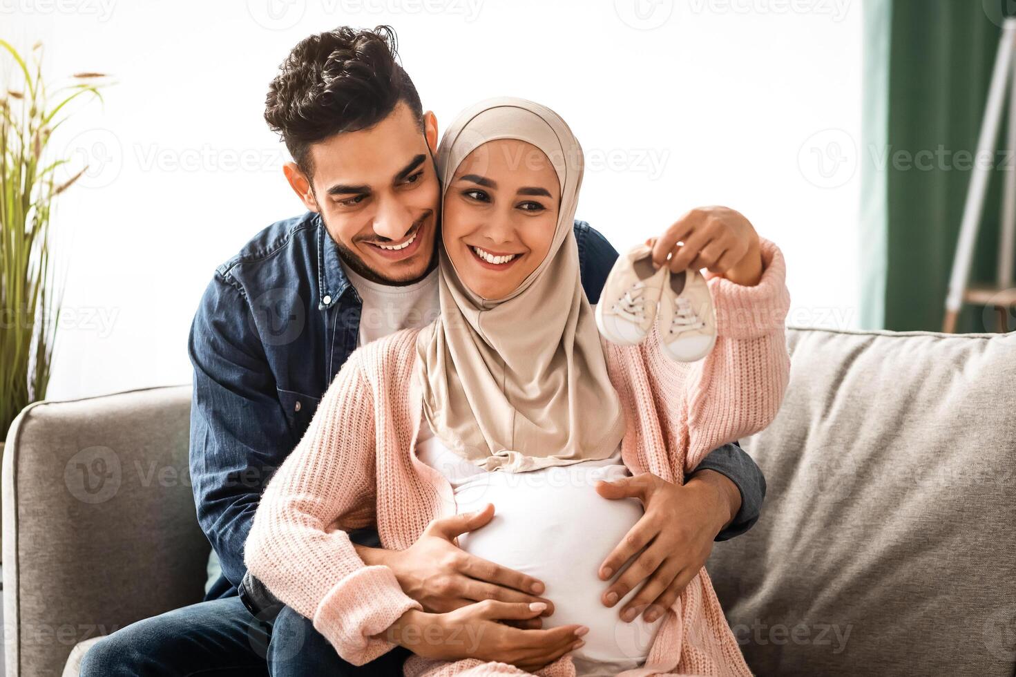 Portrait of happy pregnant muslim couple looking at tiny baby shoes while hugging on couch at home, young loving middle-eastern spouses embracing and enjoying future parenthood, free space photo