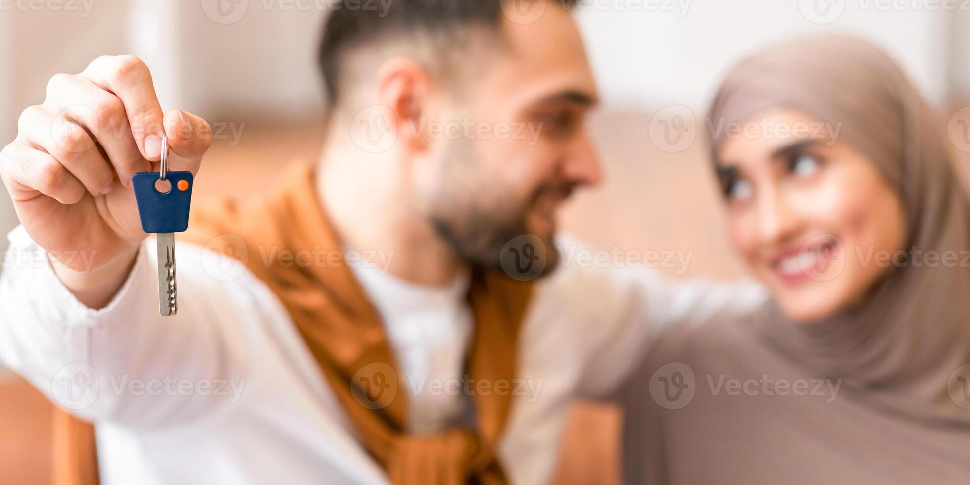 Our new home. Happy muslim couple showing new house key sitting indoors. Real estate, relocation, ownership and mortgage loan for young arab families concept. Selective focus, shallow depth photo
