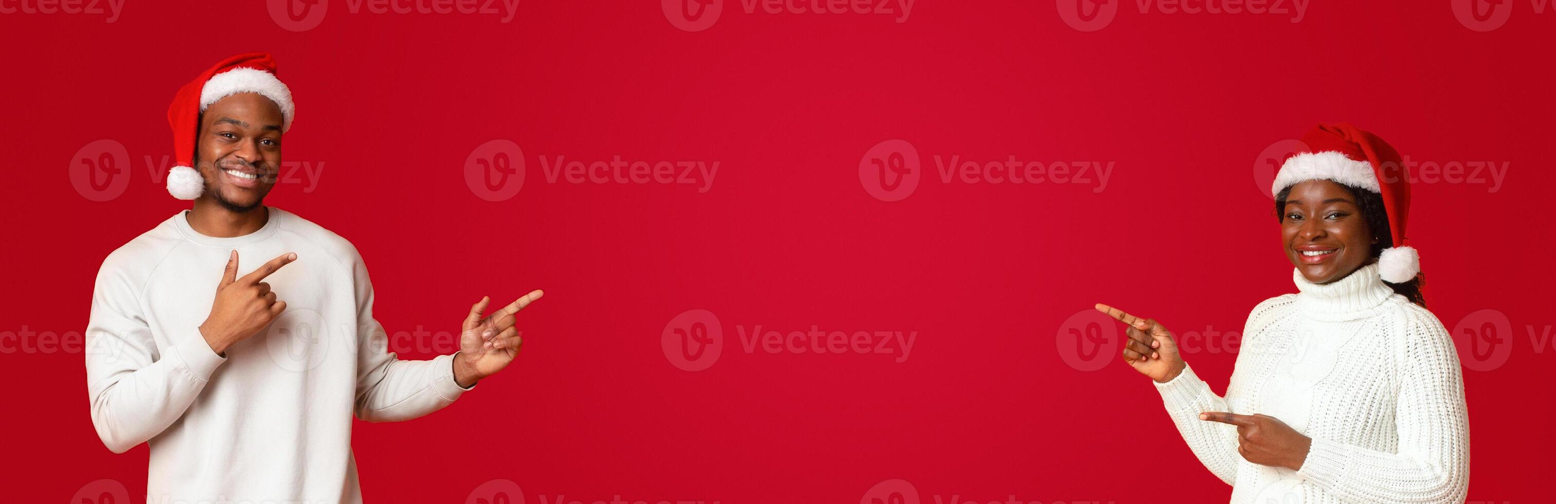 Excited african couple in santa hats pointing with fingers at free space in the middle of orange background, cheerful spouses showing empty place for your christmas offer or advertisement, panorama photo