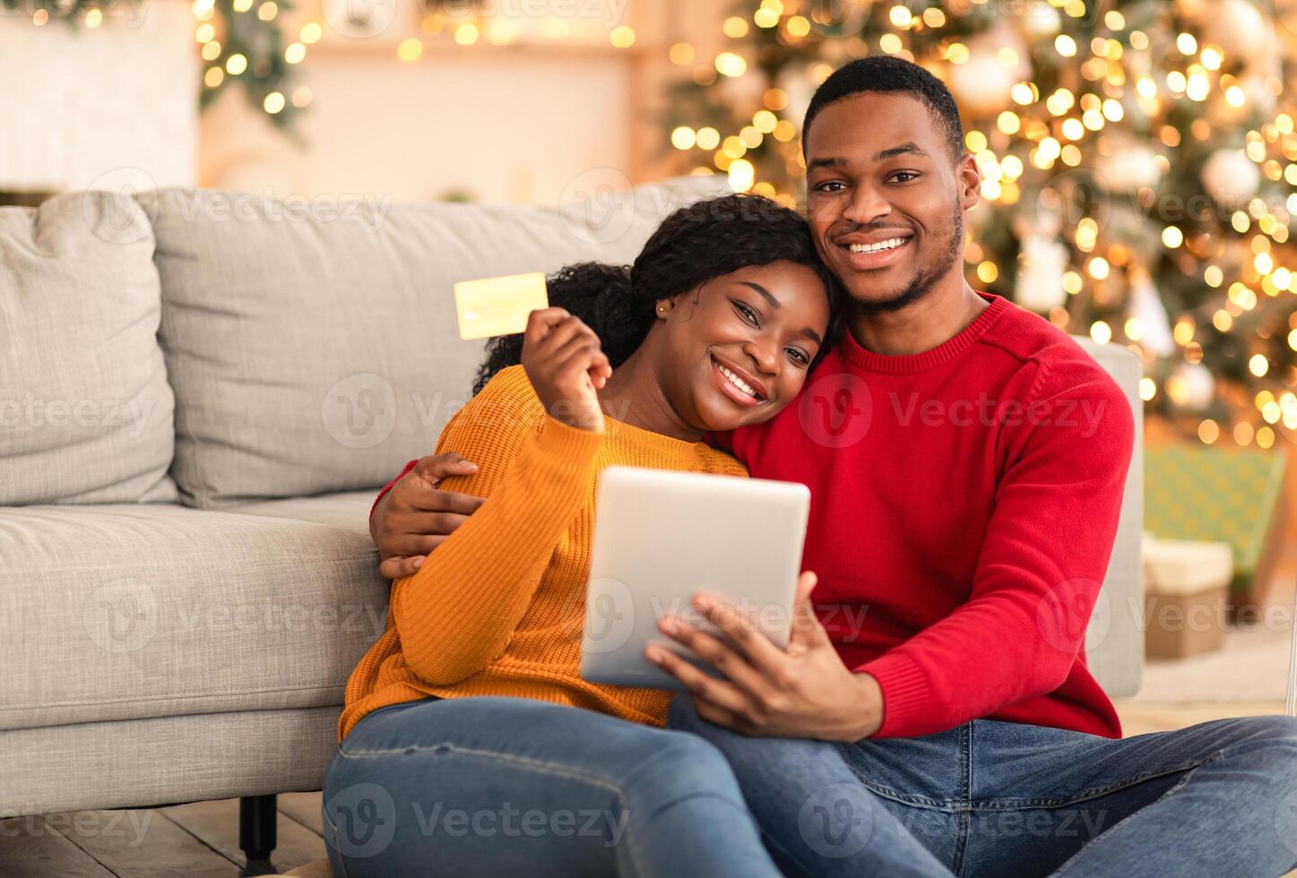 Modern new year, new normal and online during coronavirus quarantine. Millennial happy african american family use tablet and show credit card in living room with Xmas tree with glowing garlands photo