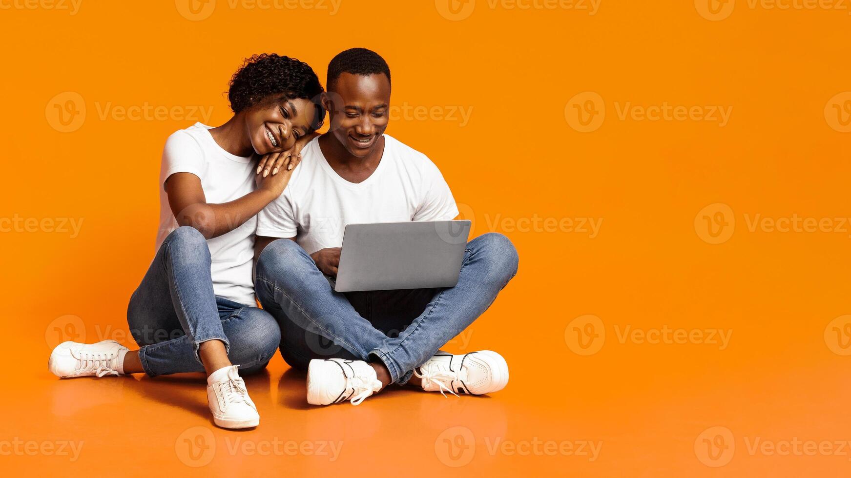 Carefree african american man and woman using new laptop together while sitting on floor, checking social media, shopping online or watching photos, orange studio background, copy space photo
