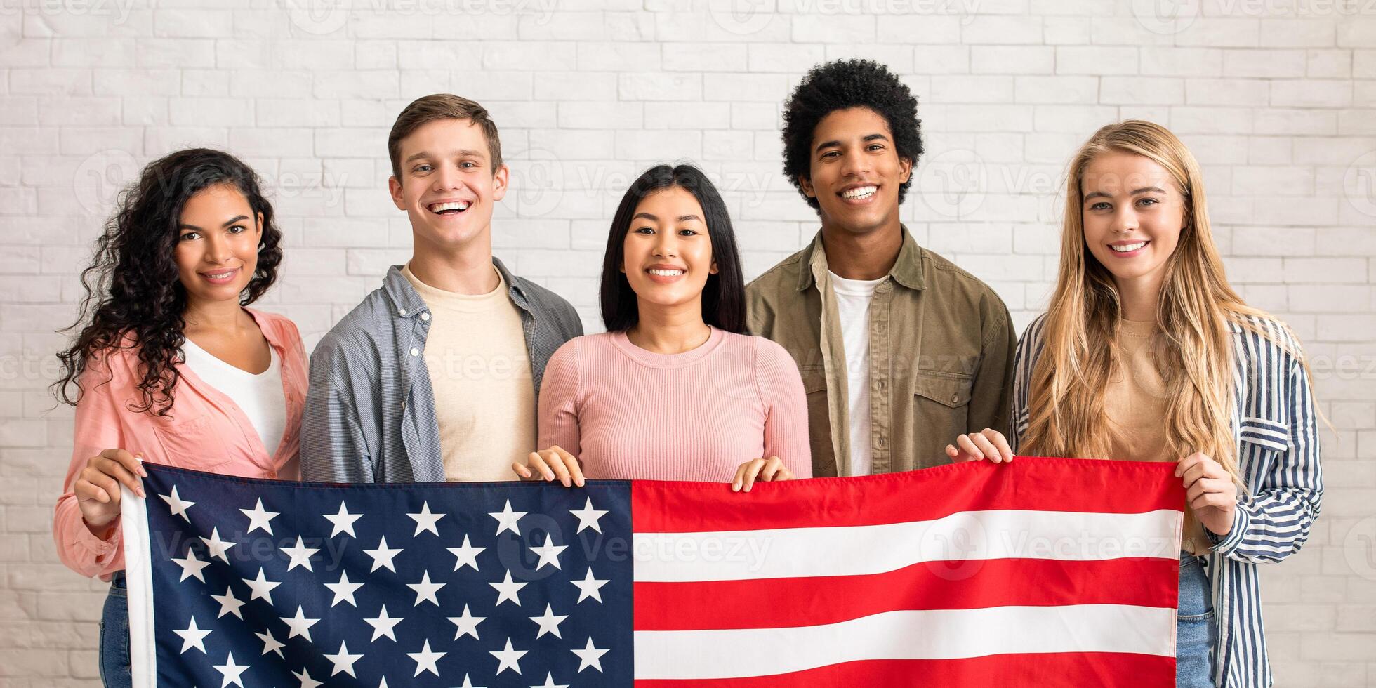 Immigration, relocation and study abroad. Cheerful young international students hold USA flag, looking at camera and ready to learn, on white brick wall background, copy space, studio shot photo