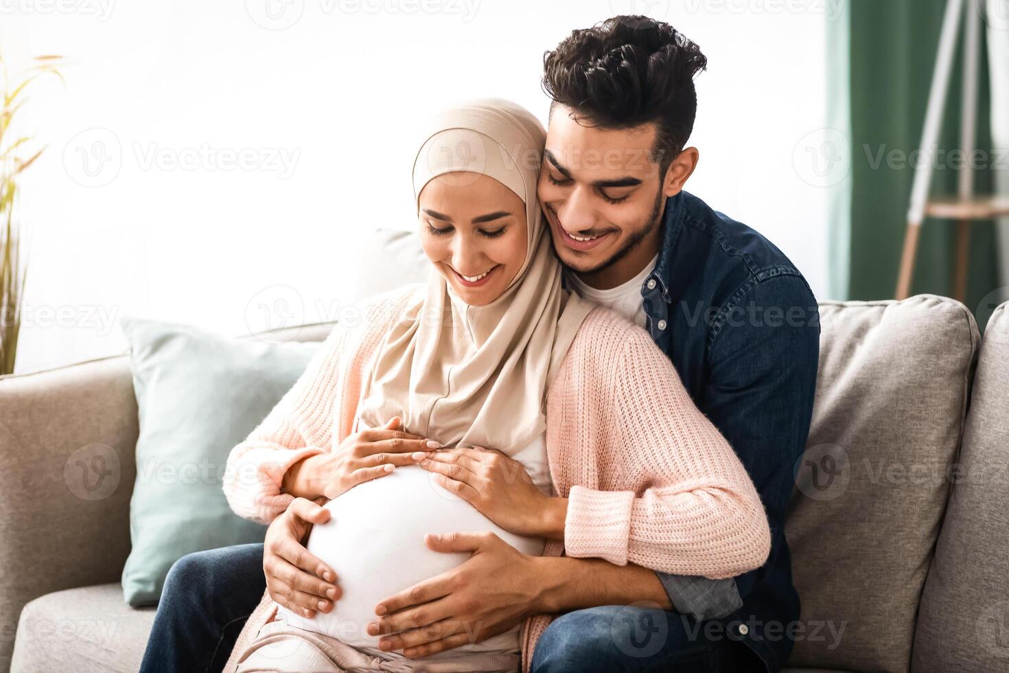 Pregnant Couple Relax. Happy Muslim Family Expecting A Baby And Bonding On Couch At Home, Cheerful Future Father Embracing His Smiling Islamic Wife In Hijab, Tenderly Touching Her Belly, Free Space photo