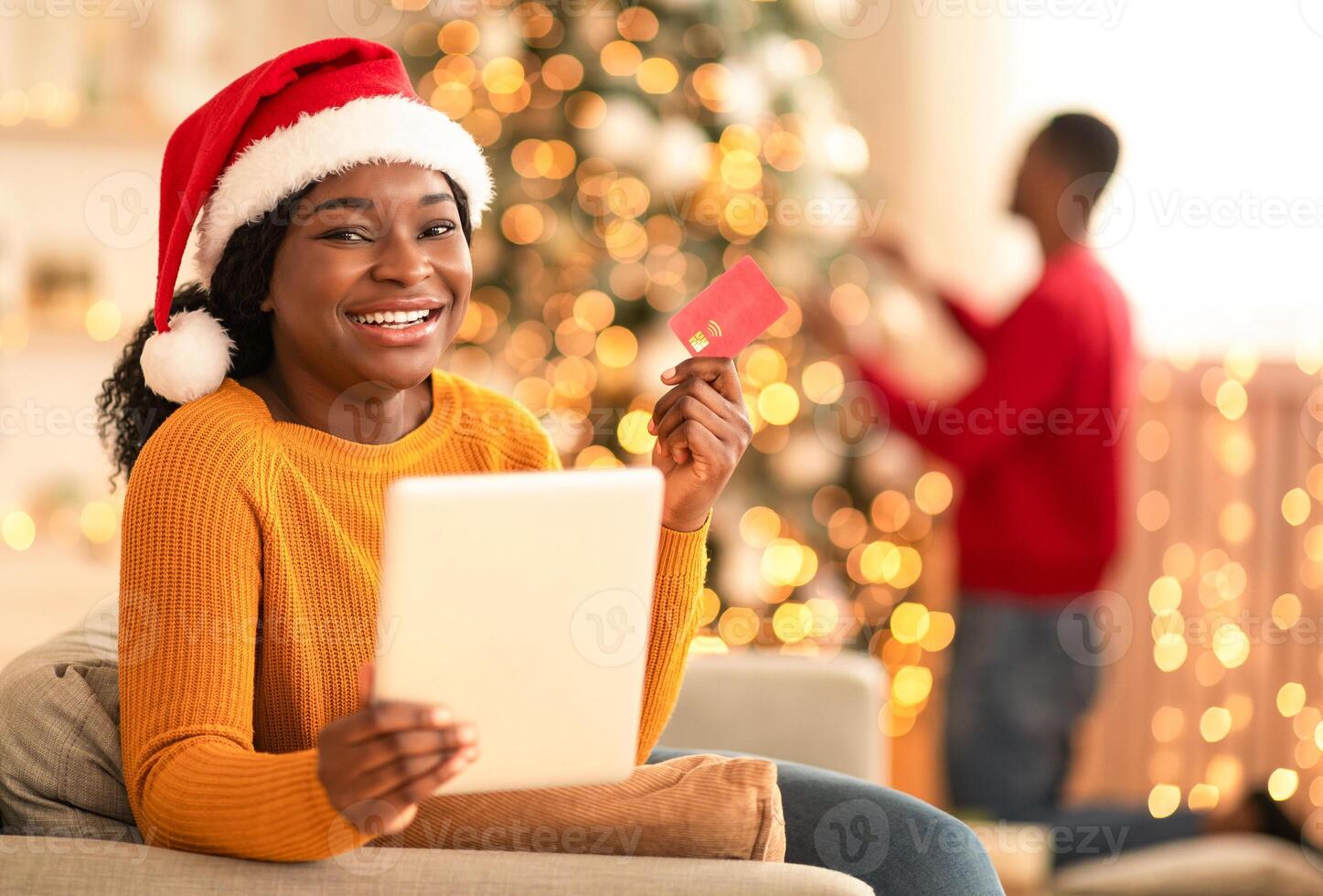 Seasonal sales and online shopping from home for holidays. Cheerful millennial african american wife in Santa hat, hold tablet and show credit card, in cozy interior, guy decorating tree with garlands photo