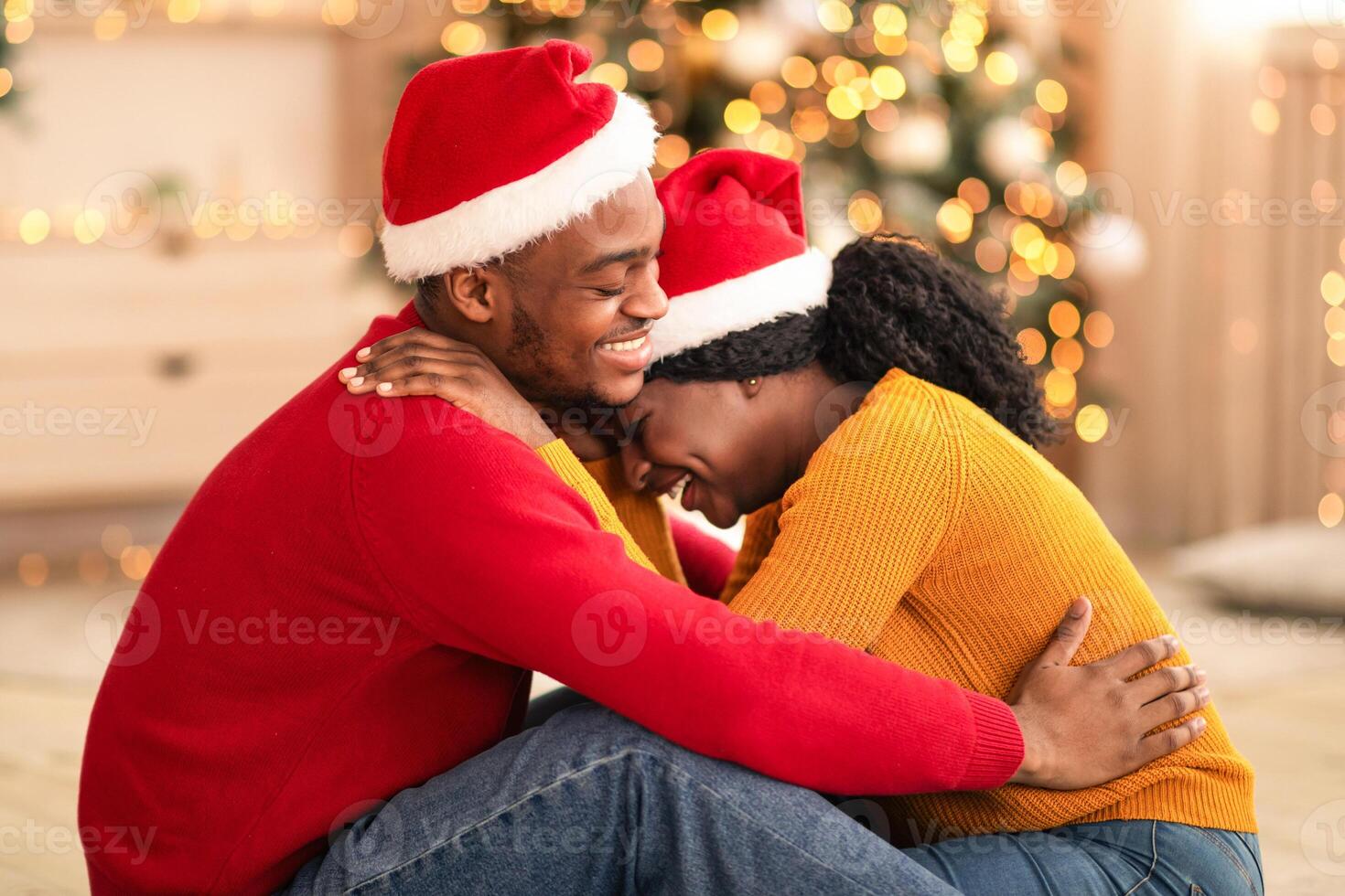 Free time together and new year at home during self-isolation. Laughing young african american guy and lady in Santa Claus hats hugging in living room with Xmas tree and decor with glowing garlands photo