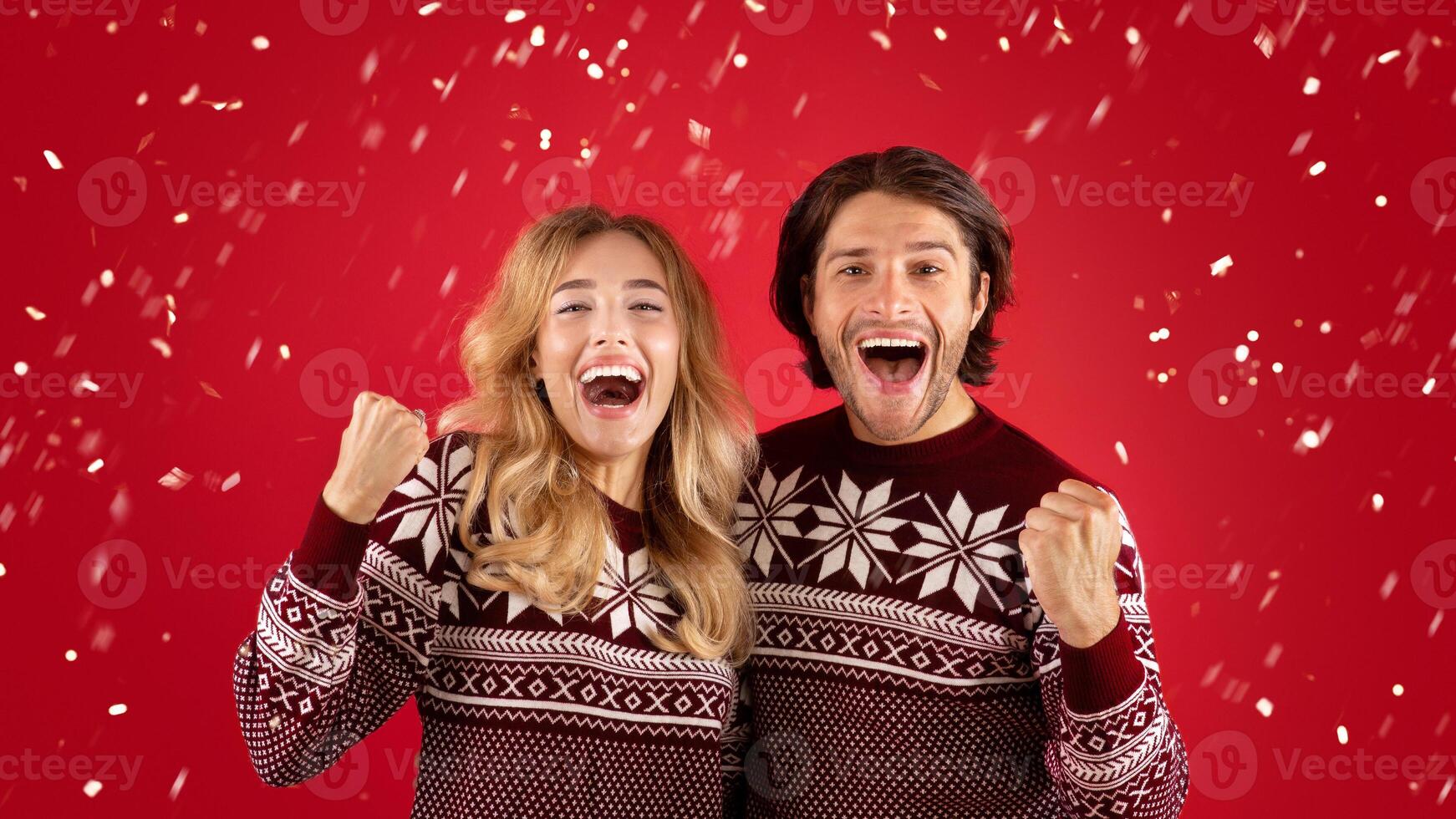 Excited young couple in Christmas sweaters screaming and making YES gesture on red background photo