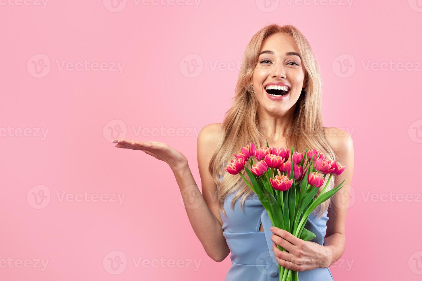 Excited blonde woman with bunch of tulips celebrating spring holiday, holding something on pink studio background, mockup for your product. Happy millennial lady posing with bouquet of flowers photo
