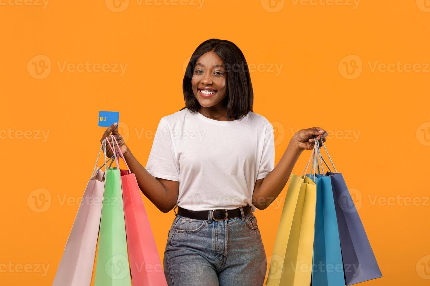 Joyful black lady with shopping bags and credit card smiling over orange studio background, empty space. African american woman shopaholic holding bunch of purchases and unlimited credit card photo