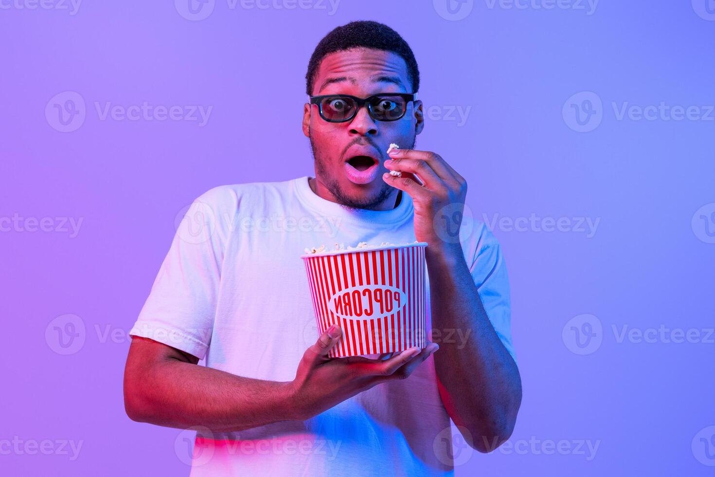 Portrait Of Shocked African American Guy In 3D Glasses Eating Popcorn From Bucket And Looking At Camera, Amazed Black Man Watching Movie In Cinema, Standing Under Neon light Over Purple Background photo