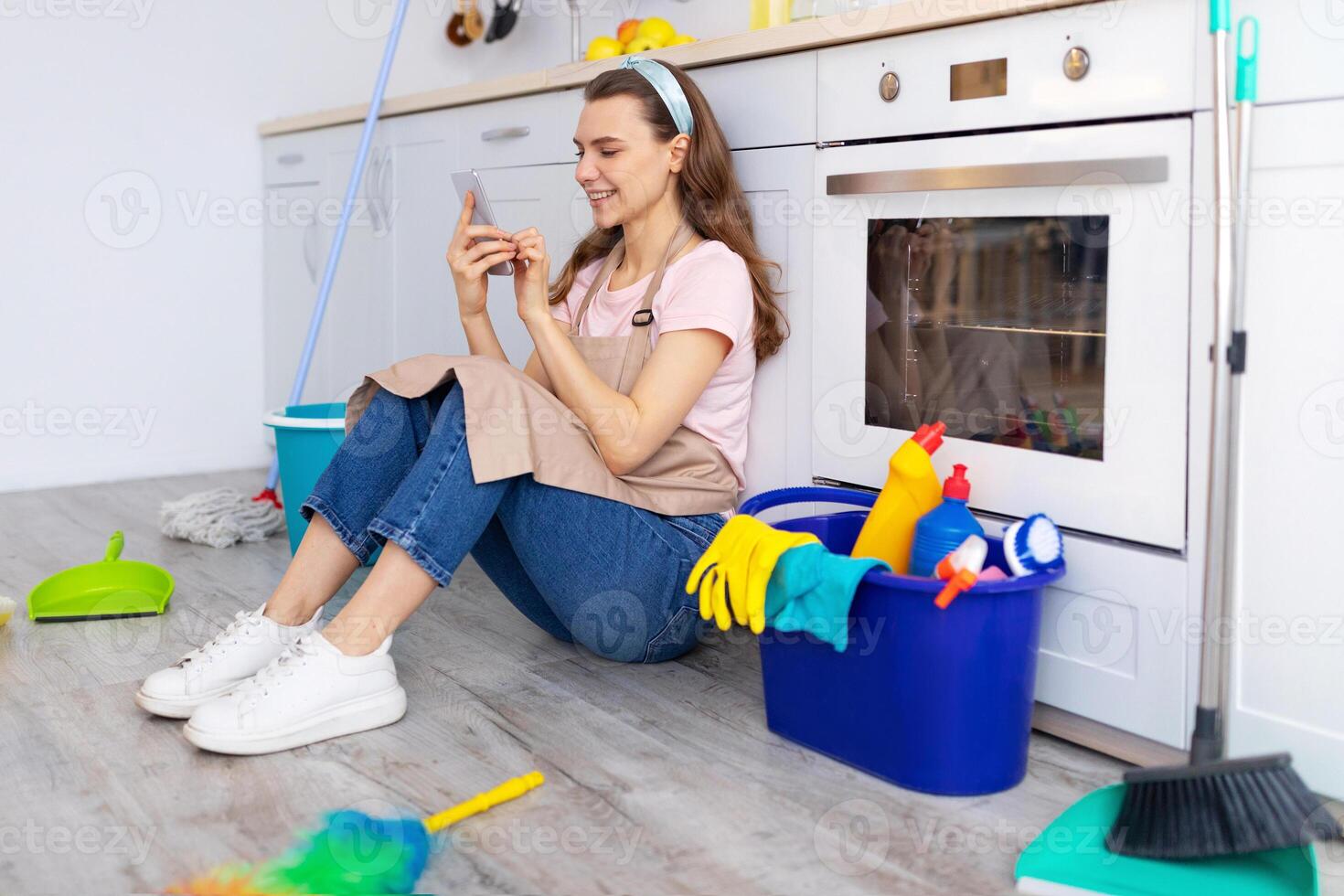 Happy young housewife taking break from home cleaning, browsing internet on smartphone while sitting on kitchen floor. Positive millennial lady checking social media during cleanup photo