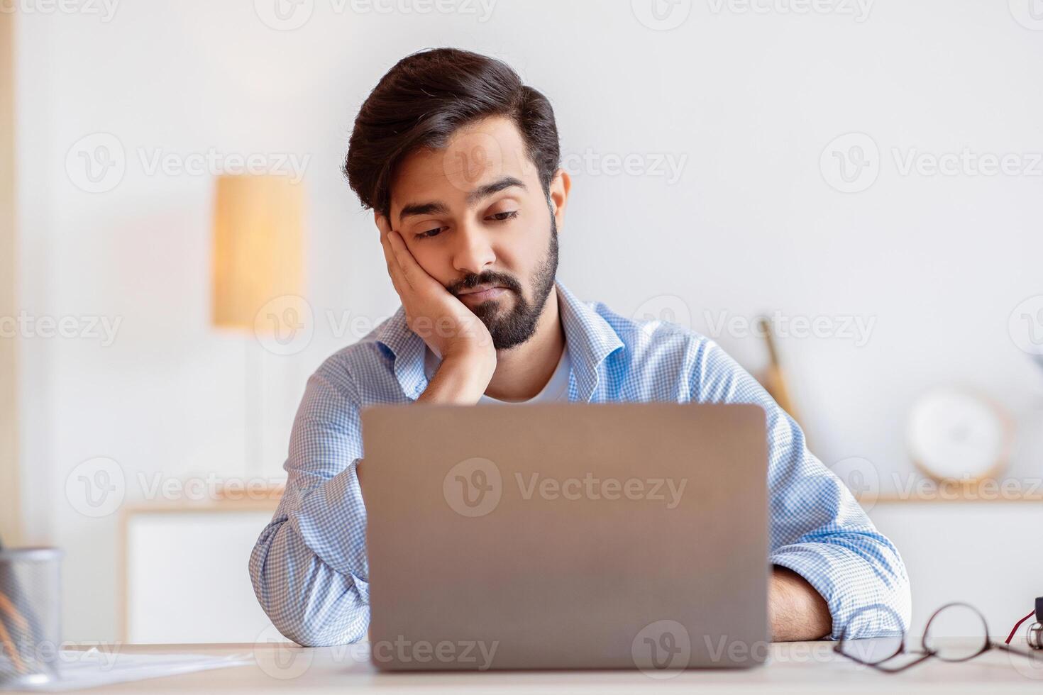 Boring Work. Young arab freelancer guy working on laptop, looking at screen with bored face expression, eastern man tired of monotonous work, sitting at desk with computer and resting head on hand photo