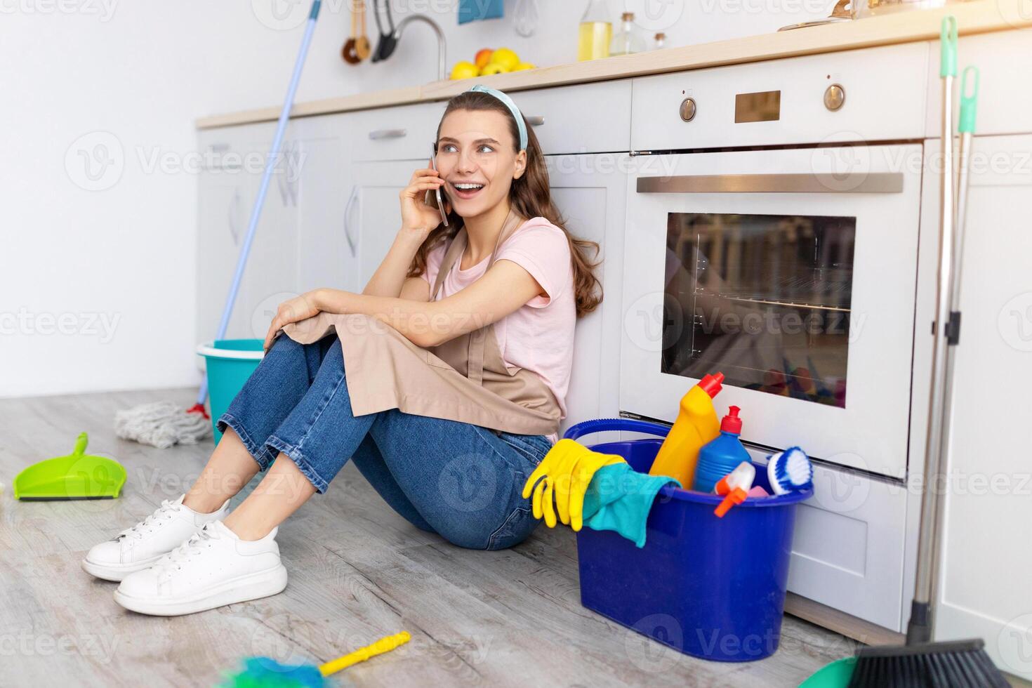 Cheerful young woman speaking on cellphone during break from housework, enjoying phone conversation with friend at kitchen. Happy housewife resting from domestic duties on floor photo