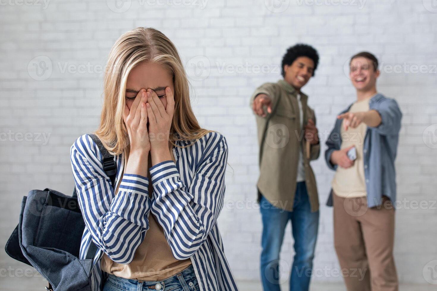 Bullying, negativity and peer problems. European and african american teens zoomers guys laugh at crying female with backpack in school or college, on white wall background, studio shot, empty space photo