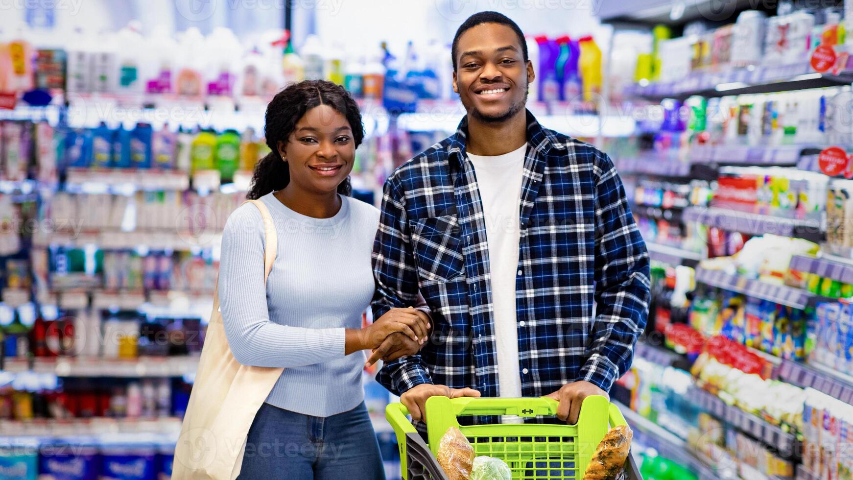 Portrait of smiling black couple with shopping cart full of products looking at camera in modern supermarket. Joyful young African American family purchasing groceries together at mall photo