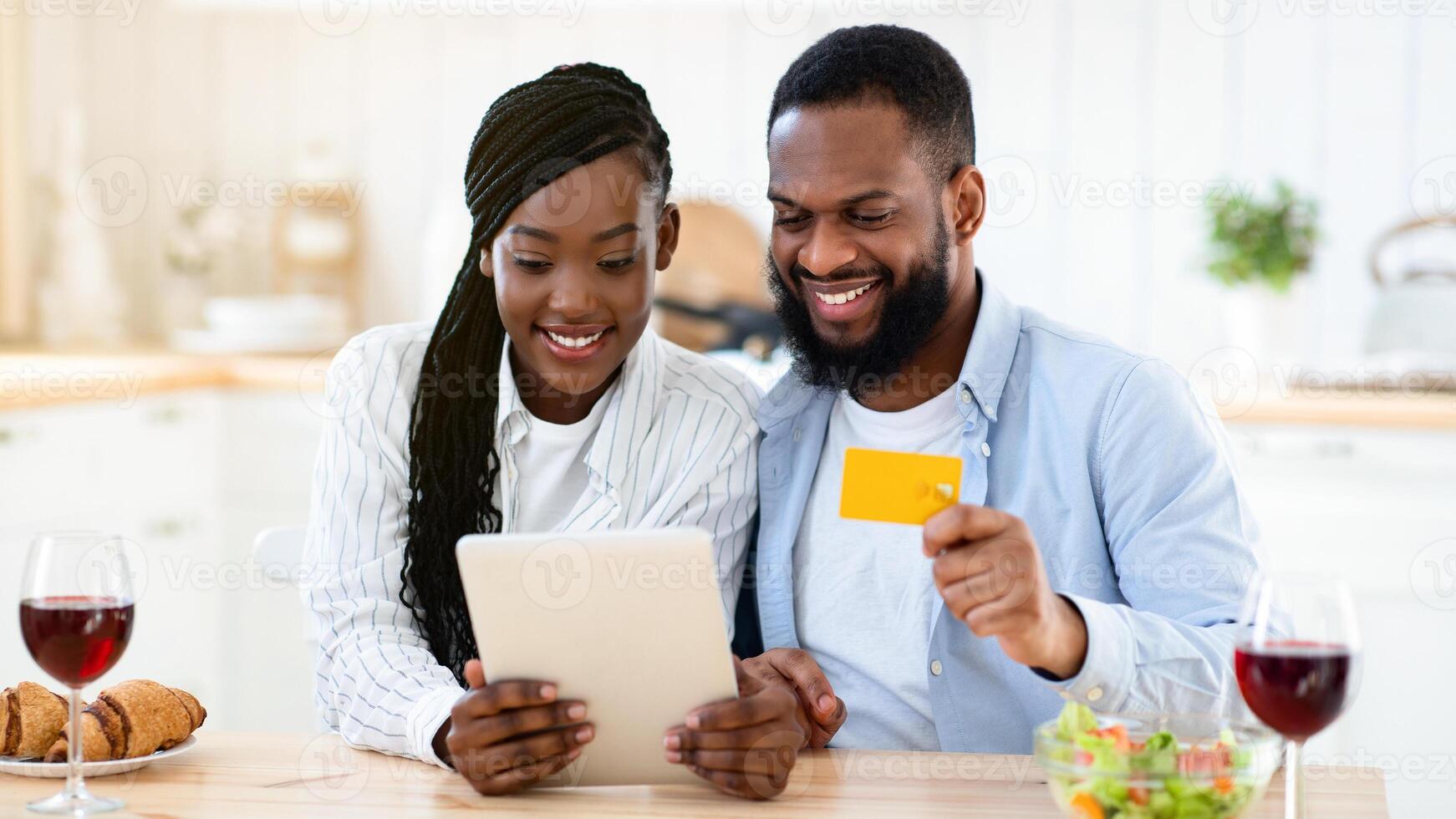 Cheerful Black Spouses Ordering Food Online With Digital Tablet And Credit Card, Sitting At Table In Kitchen, Purchasing Grocery Delivery, Enjoying Modern Technologies For Daily Life, Closeup photo