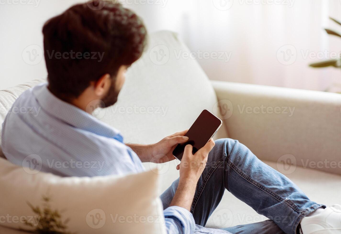 Unrecognizable Man Using Smartphone With Blank Black Screen While Relaxing On Couch At Home. Mockup Image With Bearded Man Messaging Or Shopping Online On Cellphone, Resting In Living Room, Copy Space photo