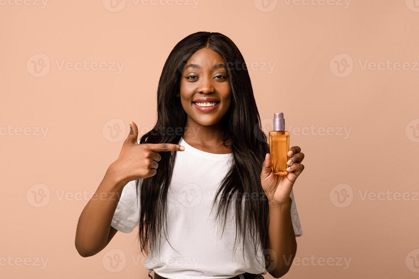 Pretty young black woman with long beautiful hair pointing at bottle with hair product, serum or oil, beige studio background, copy space. Hair care, healthy and beautiful hair concept photo