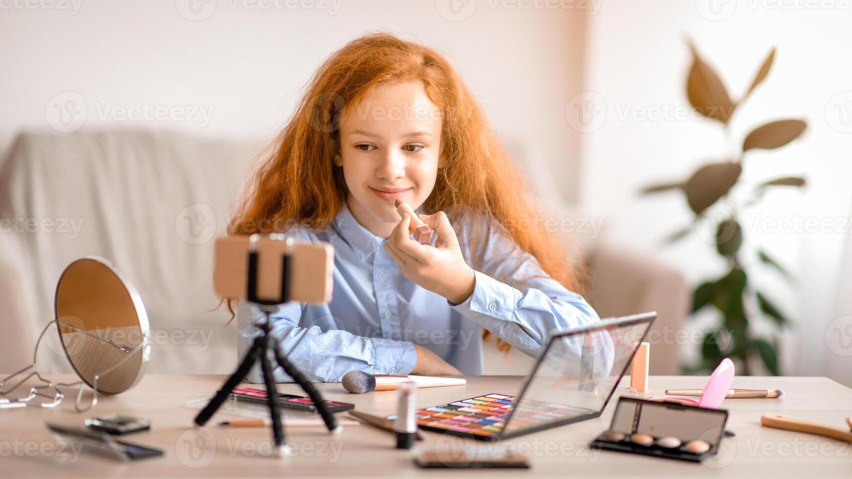 Beauty Blog For Teenagers. Young beautiful blogger doing makeup, recording cosmetics product review, applying lipstick and looking at camera. Girl filming vlog for her channel with cellphone on tripod photo