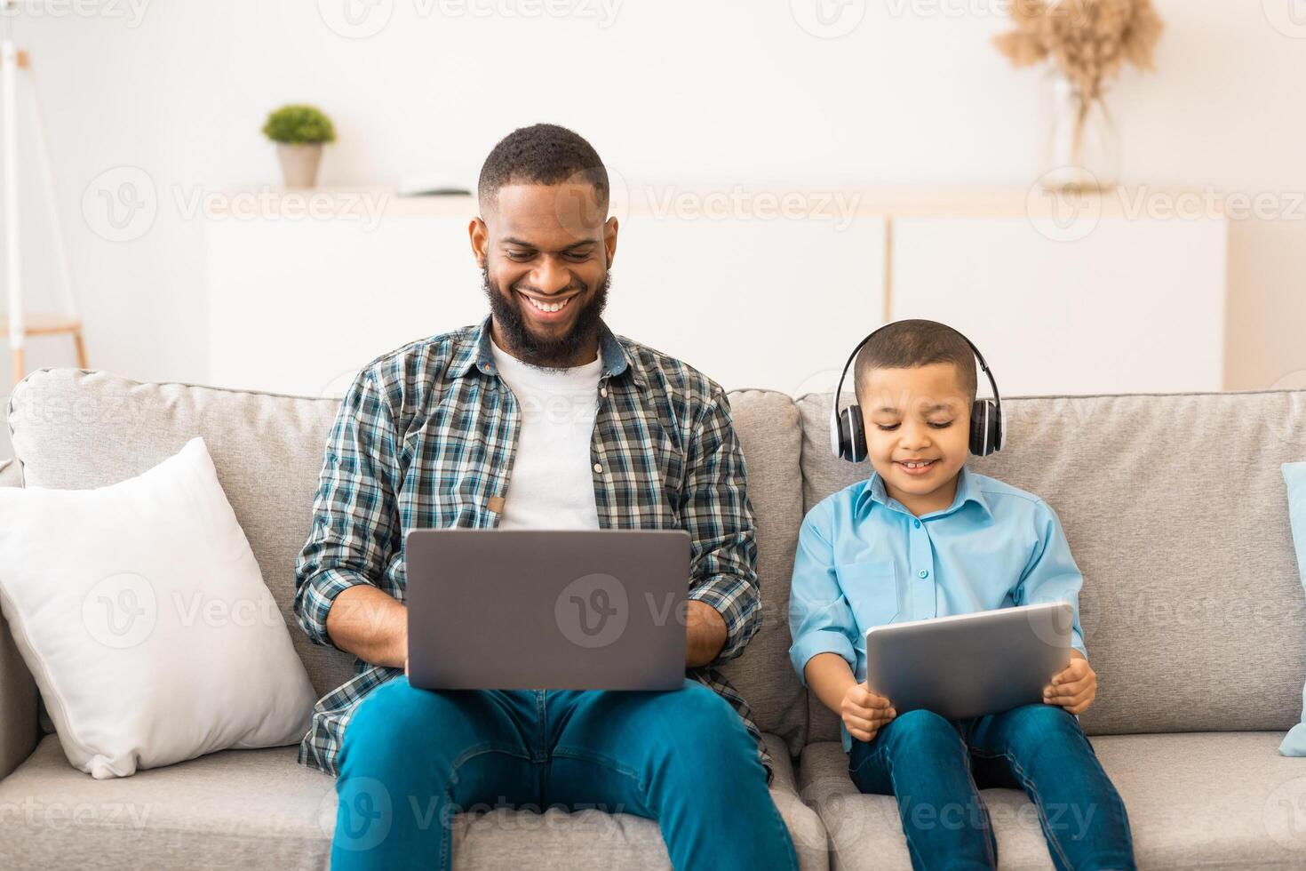 Black Father And Little Boy Using Computers Together Browsing Internet On Laptop And Digital Tablet Wearing Wireless Headphones Sitting On Sofa At Home. Family Gadgets Concept photo