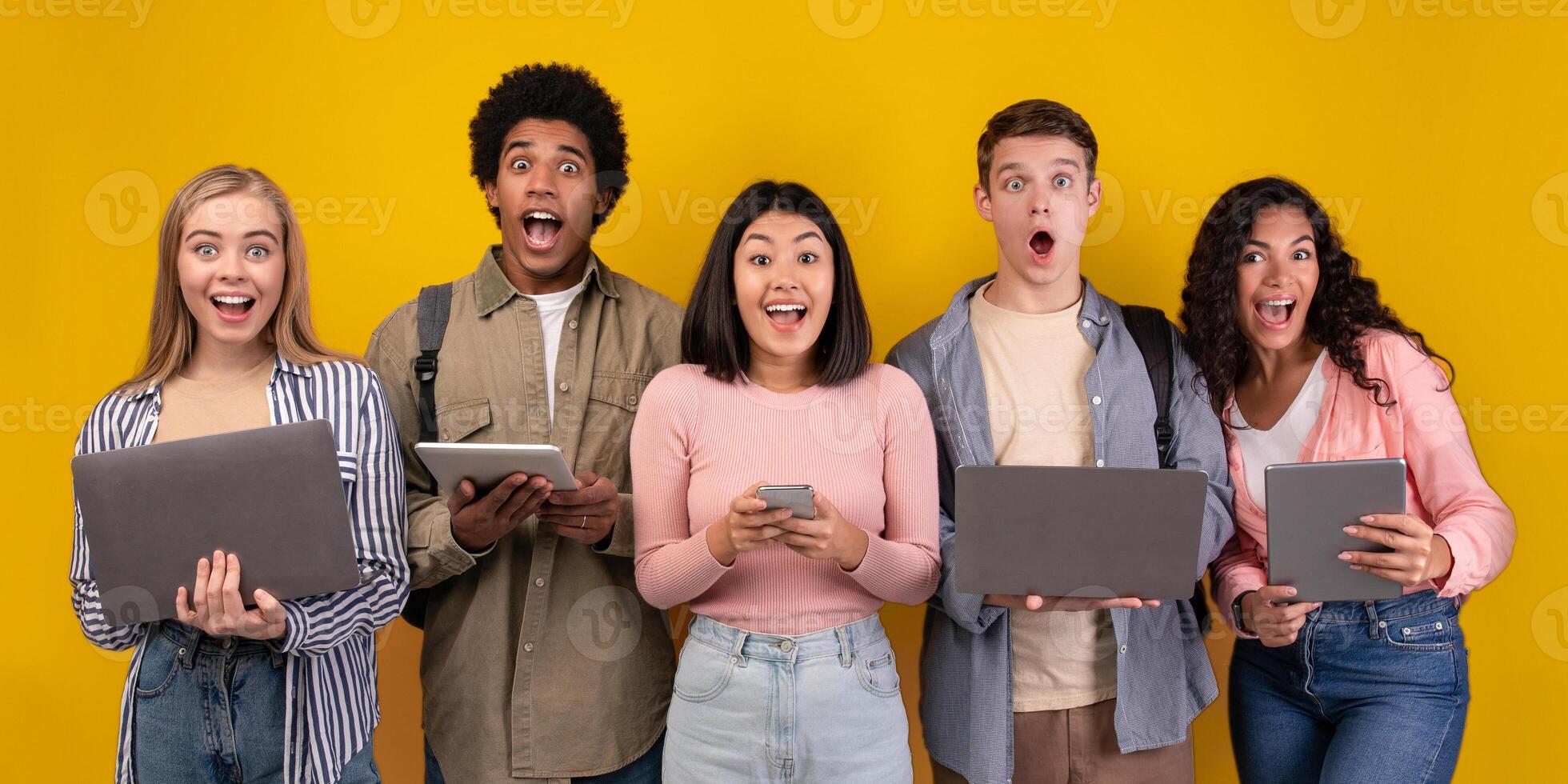 Gossip, shocking news, study with online technology and teamwork. Surprised teens multiracial students learning, using digital gadgets, isolated on orange background, copy space, studio shot photo