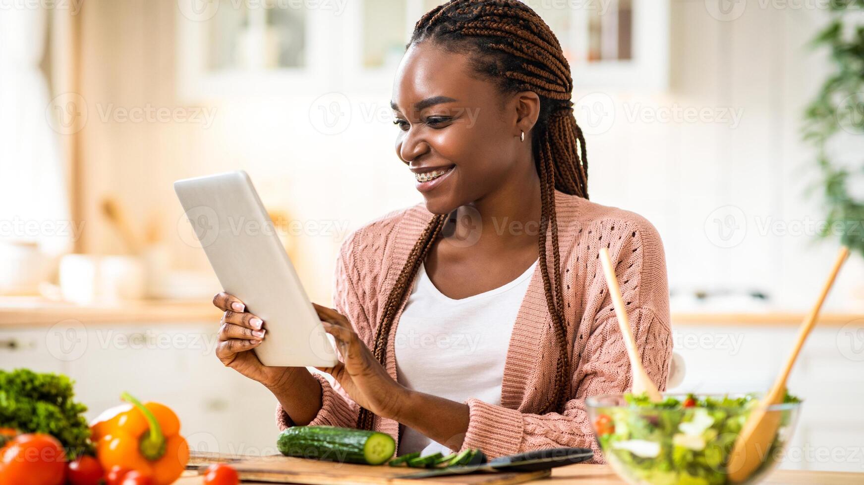 Online Recipe. Cheerful african american woman using digital tablet in kitchen while cooking healthy lunch, smiling black female checking ingredients in internet or reading culinary blog, free space photo