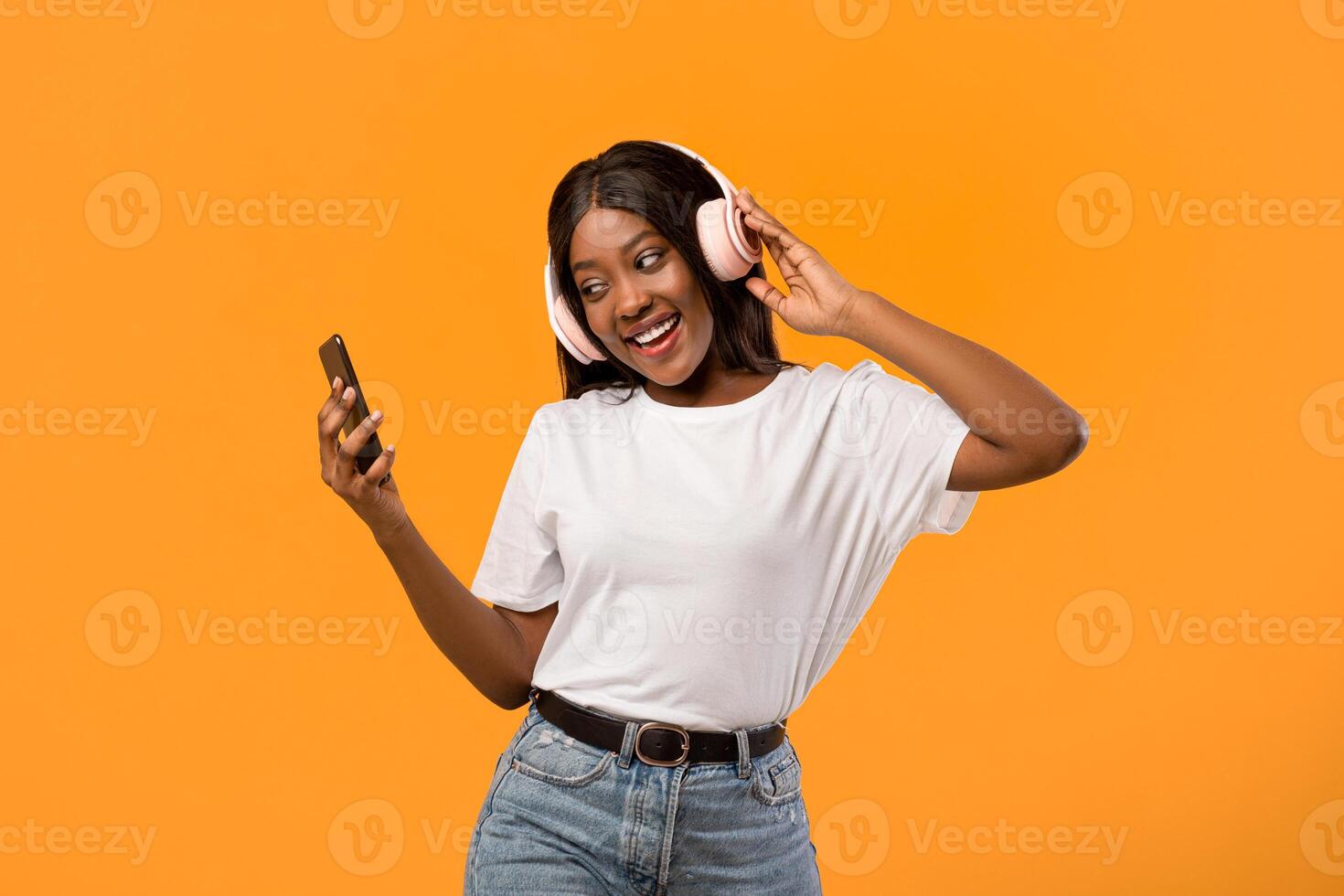 Joyful young african american lady listening to music over orange studio background, using wireless headphones and smartphone. Smiling black woman enjoying newest stereo headset and mobile music app photo