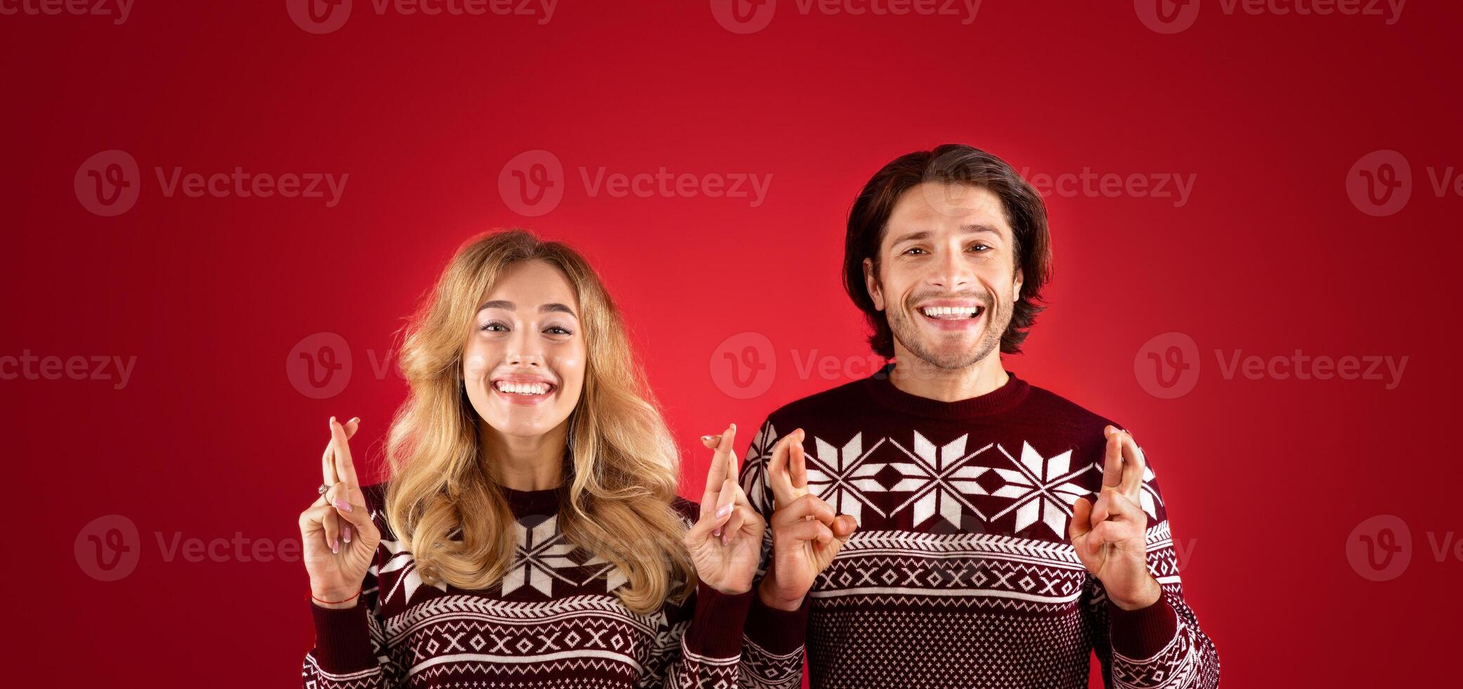 Making Christmas wish. Beautiful young couple in winter sweaters crossing fingers for luck on red background photo