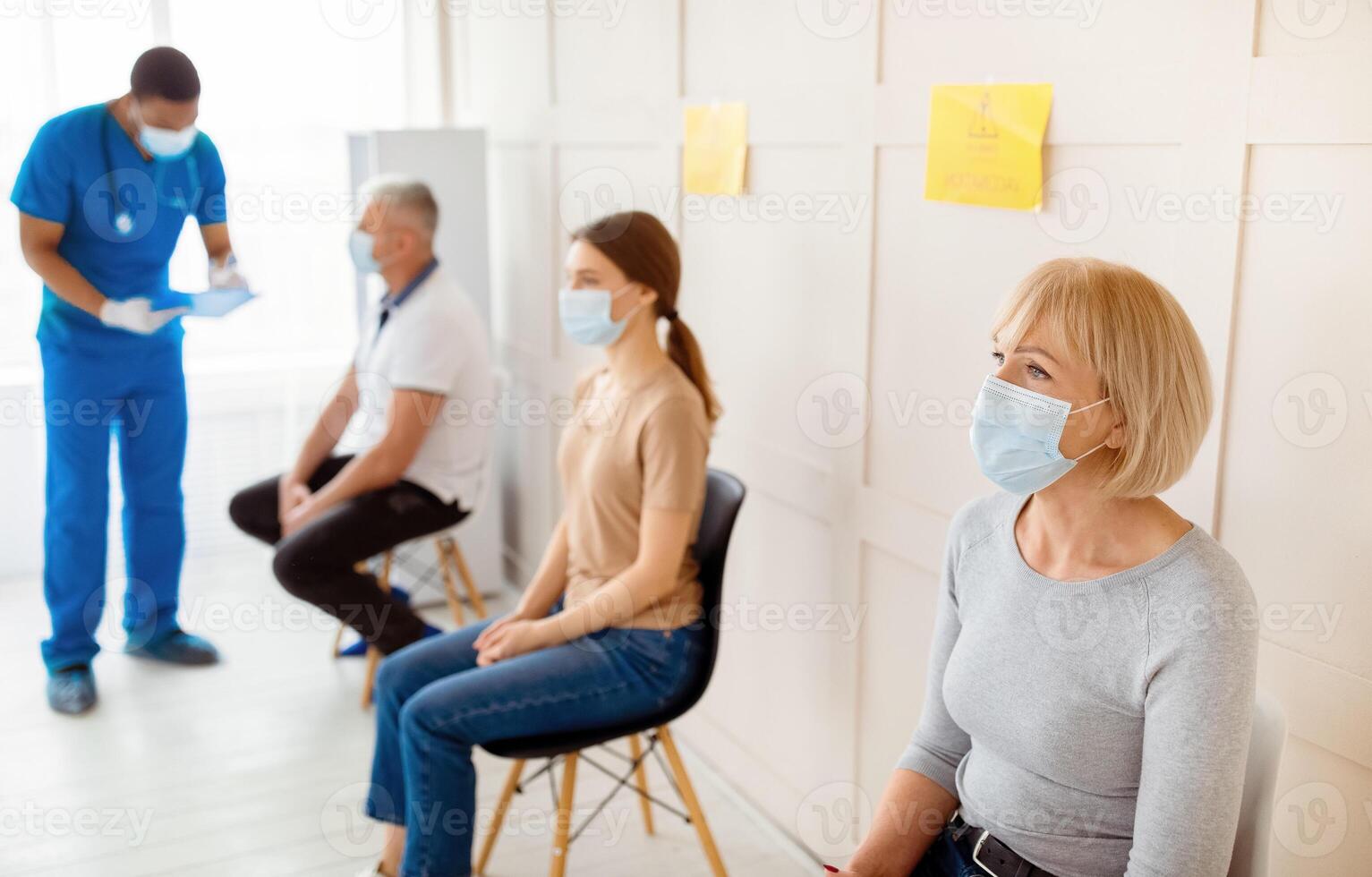 Mature woman and other patients in face masks waiting in queue for vaccination against covid-19 at clinic, communicating to doctor, taking part in anti coronavirus campaign, copy space photo