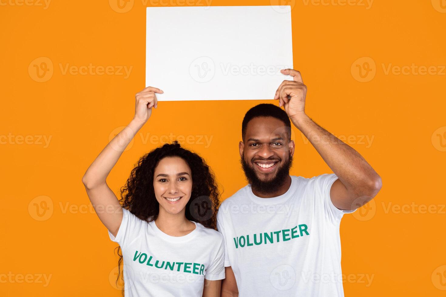 Couple Of Multiethnic Volunteers Holding Empty White Paper Board Above Heads Advertising Your Text Posing Over Yellow Studio Background. Volunteering And Charity Banner Advertisement. Mockup photo