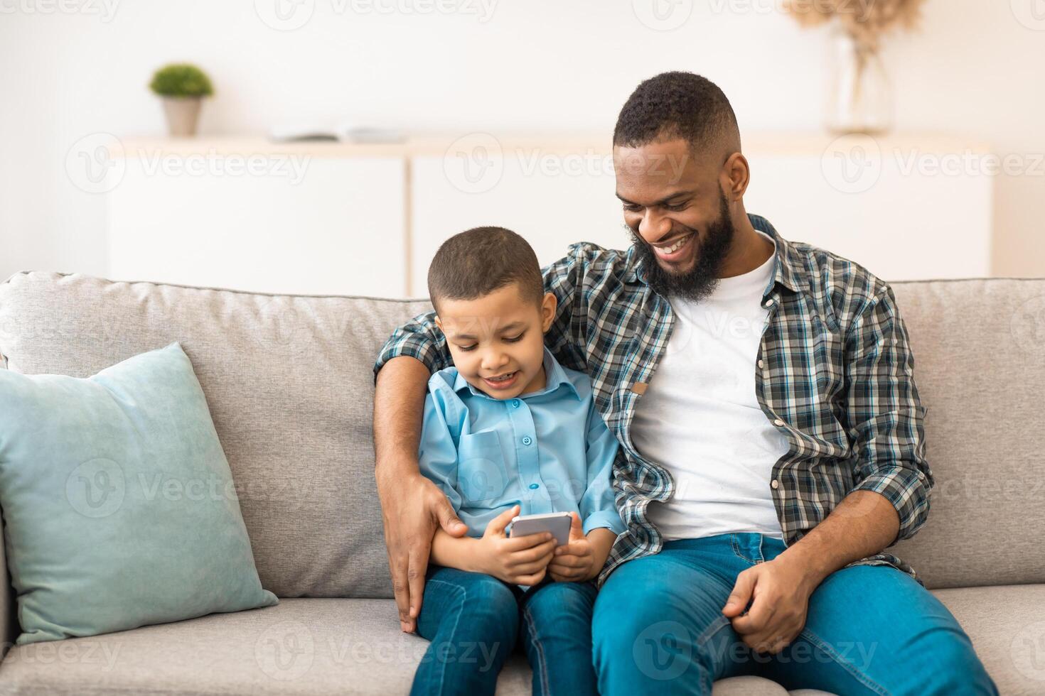 Black Boy And His Dad Using Mobile Phone Playing Games Online Sitting Together On Couch And Browsing Internet In Living Room Indoor. New Application For Smartphone, Family And Gadgets Concept photo