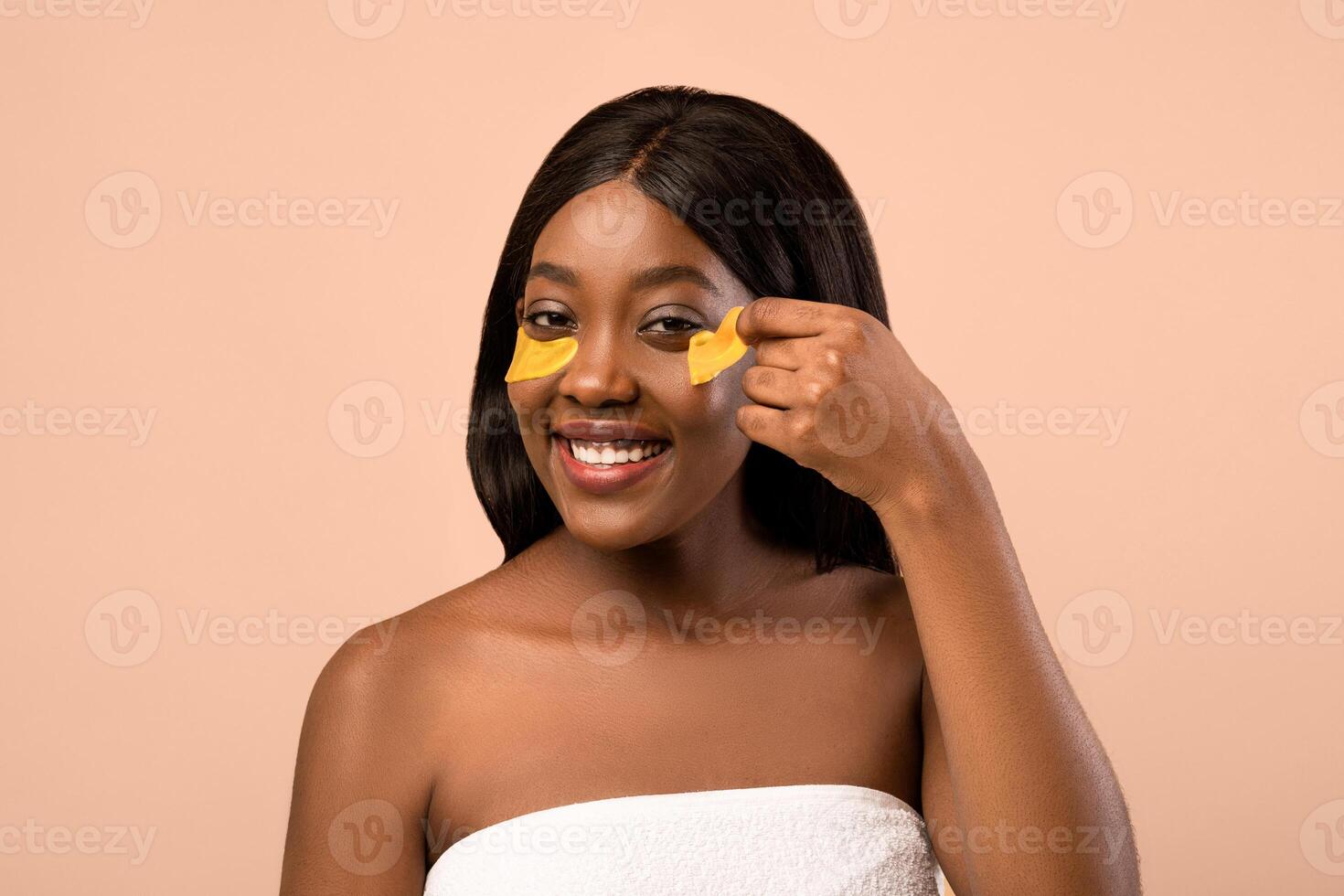 Closeup of smiling beautiful young black lady removing golden hydrogel eye patches over beige studio background, copy space. Anti-aging, against lines cosmetic tools, wrinkles eye care concept photo