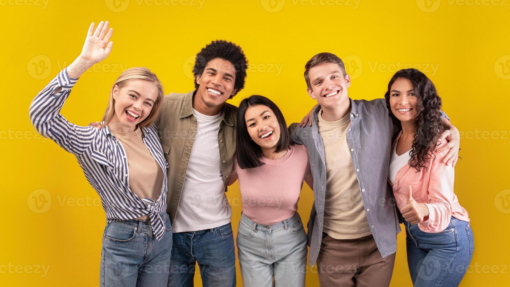 Education at university or college abroad. Cheerful young multiracial international students have fun, laughing, hugging and waving to camera isolated on orange background, copy space, studio shot photo