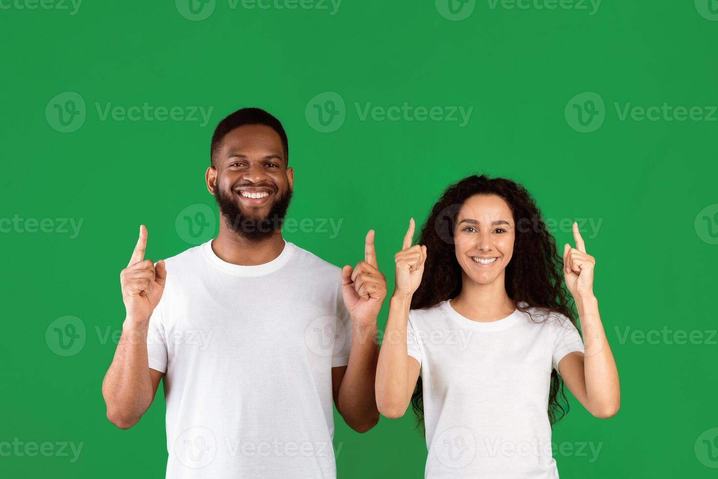 Diverse Couple Pointing Fingers Up Posing Standing In Studio Over Green Background, Smiling To Camera. Multiethnic Man And Woman Showing Something Above Their Heads. Look Upward, Advertisement Banner photo