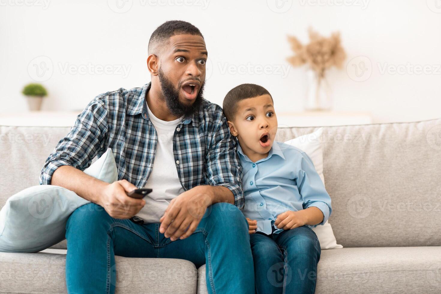 Television Programming Problem. Shocked African Father And Son Watching Questionable Content On TV Together Sitting On Couch With Opened Mouth At Home. Parental Control Concept photo