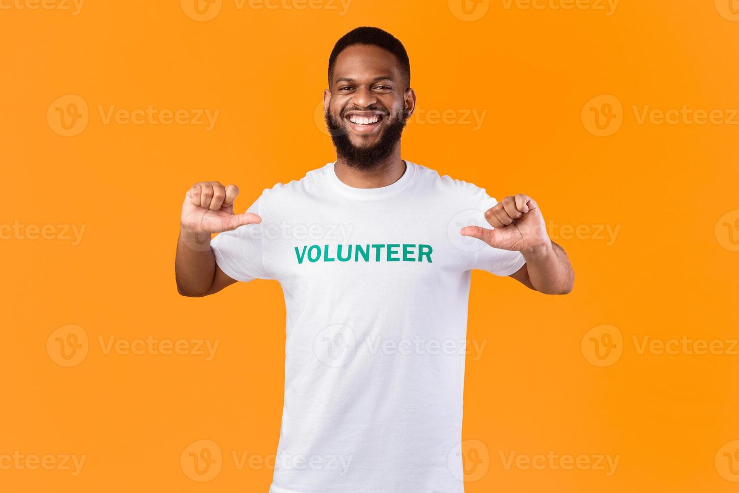 I'm a volunteer. Cheerful black activist guy pointing fingers at himself and volunteer inscription on his t-shirt posing smiling to camera on yellow studio background. Volunteering work concept photo