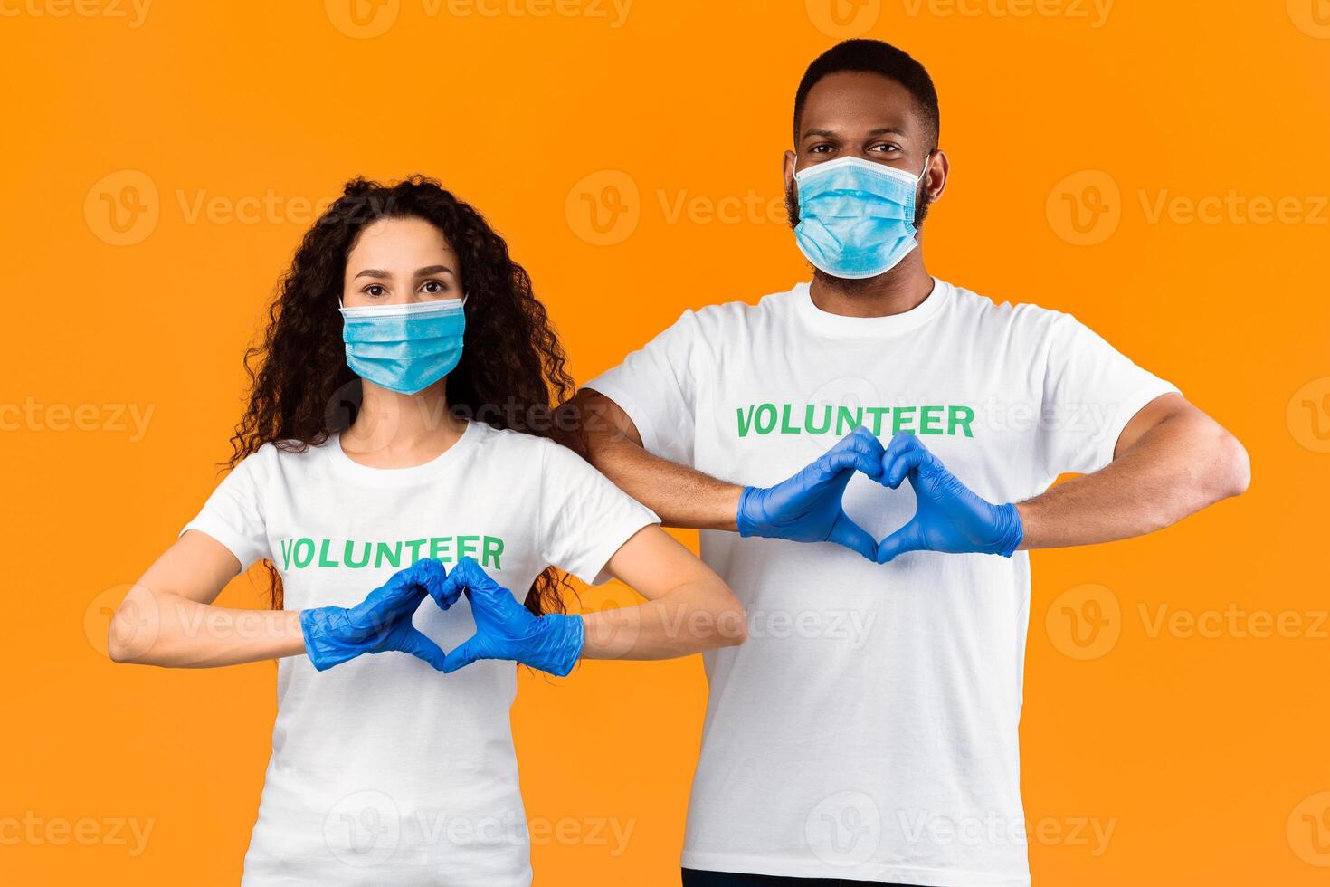 Two Multiethnic Volunteers Gesturing Heart Shape Wearing Protective Face Masks And Medical Rubber Gloves For Coronavirus Protection Posing On Yellow Studio Background. Kindness And Charity Concept photo