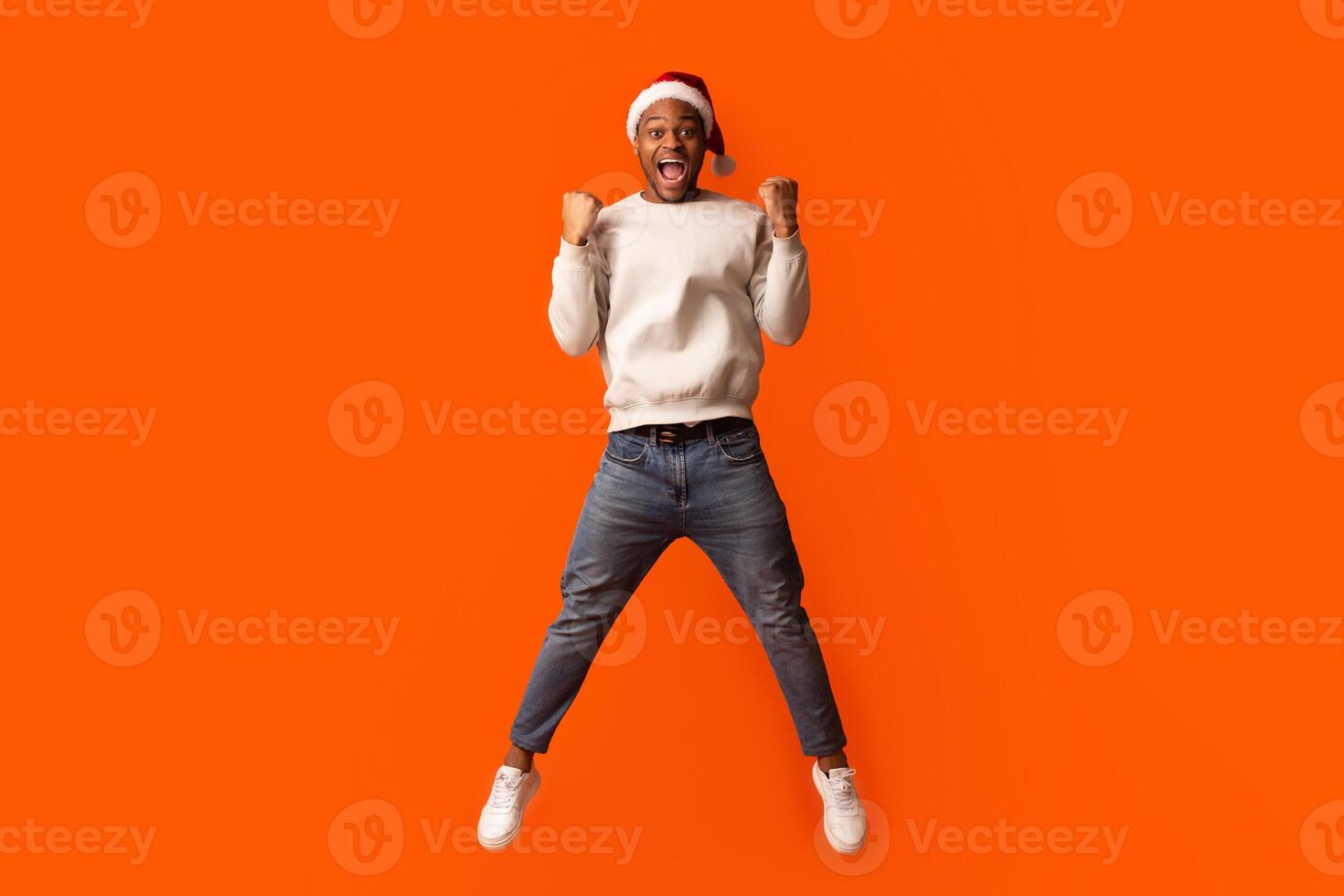 Overjoyed Black Guy In Santa Hat Celebrating Success, Jumping Over Orange Background With Raised Fists, Euphoris African American Man Enjoying Christmas Offers And Seasonal Sales, Full Length Shot photo