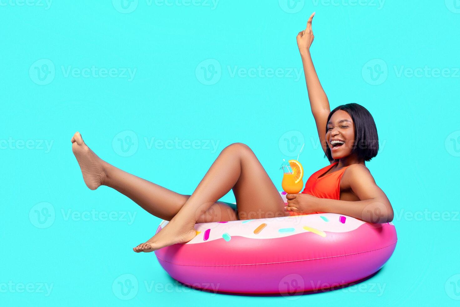 Cool black lady sitting on inflatable ring, having fun, enjoying summer cocktail on blue background, full length. Cheerful young woman having beach party, chilling and screaming in excitement photo