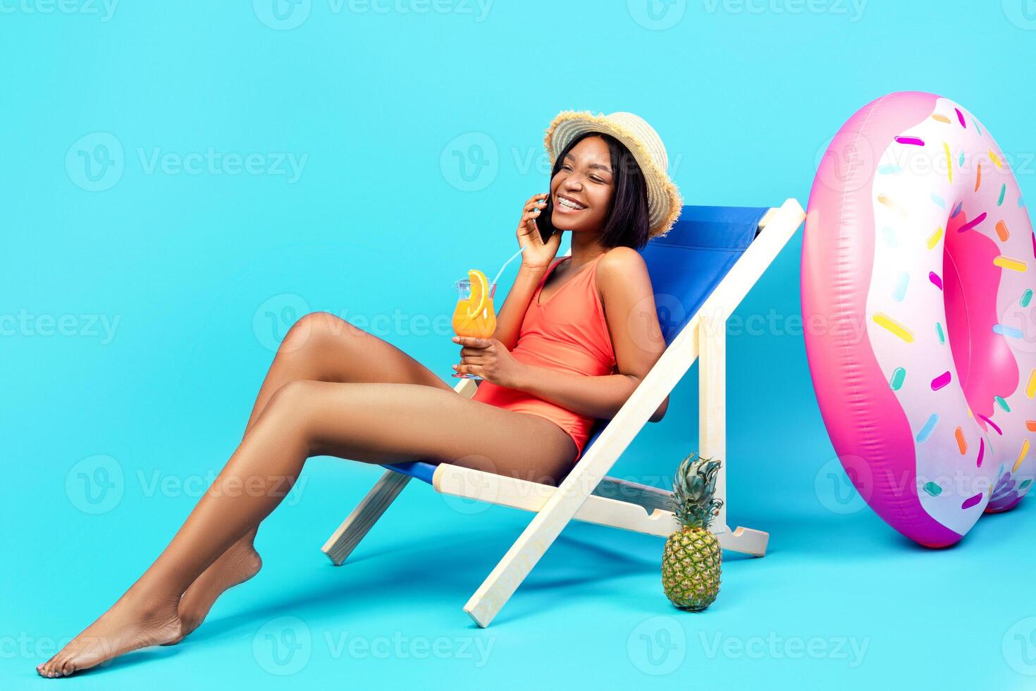 Sweet black woman in swimsuit enjoying summer cocktail, speaking on smartphone over blue studio background, panorama with copy space. Young lady having phone conversation and refreshing drink photo