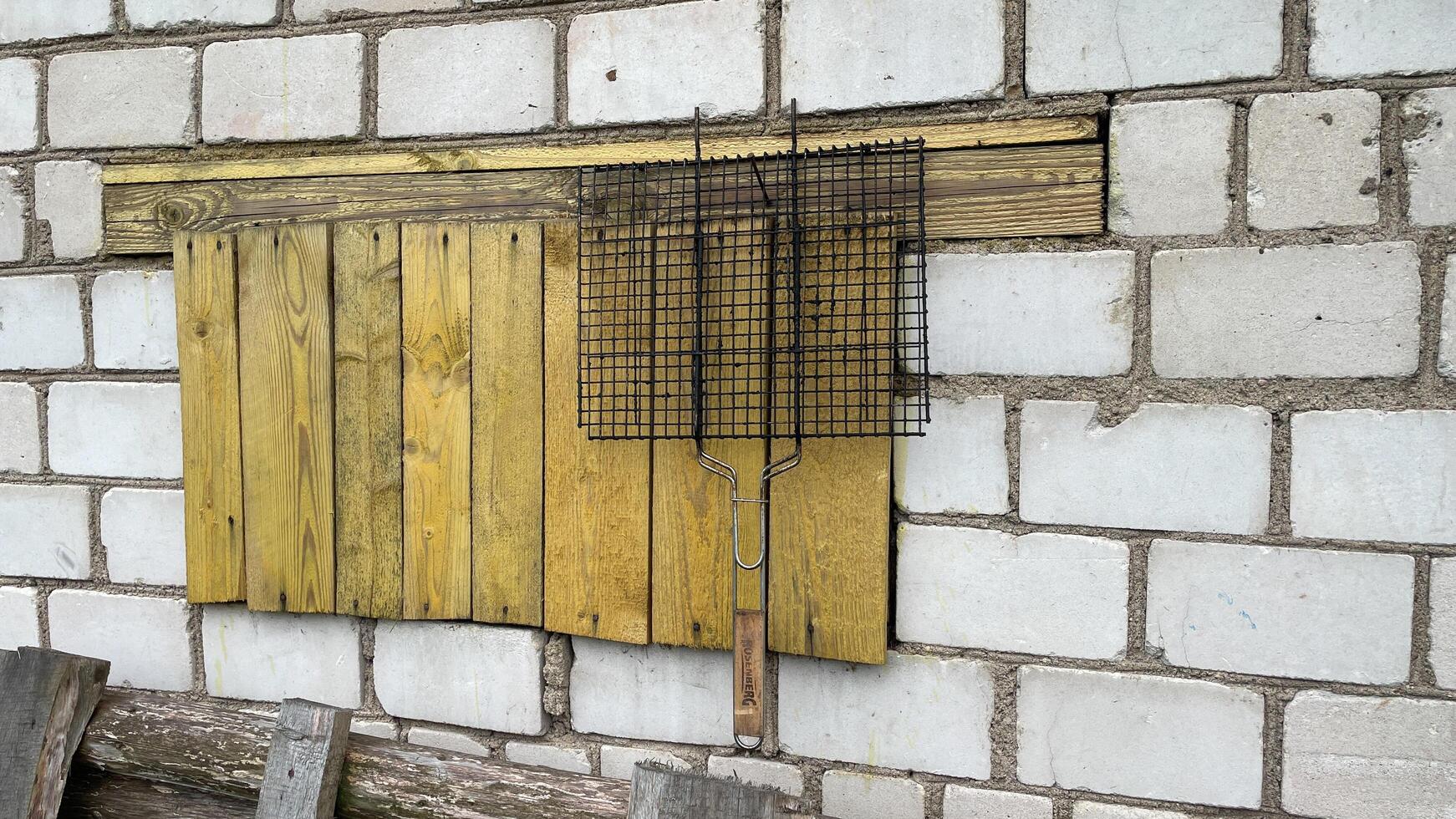 grill for roasting meat hangs on a nail in the yard of the house, against the background of a white brick wall, an old wooden stepladder stands below, yellow planks photo