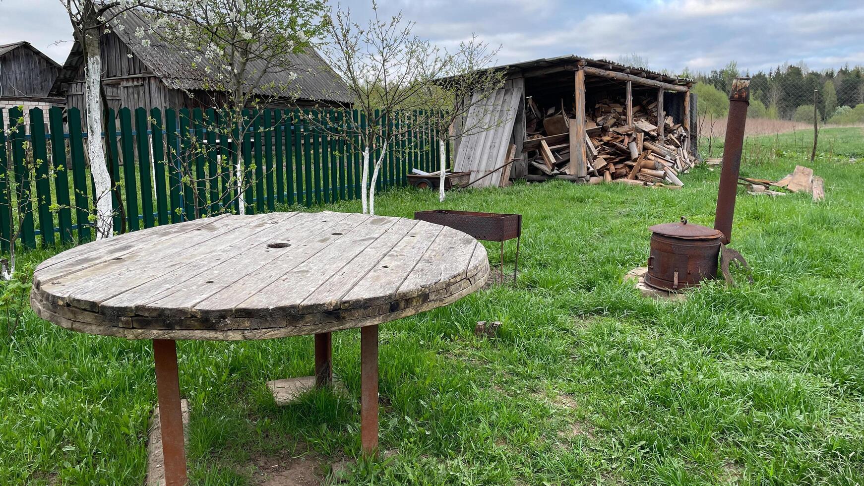 in the backyard of the house there is an old wooden shed with boards, a round wooden table for guests, a homemade stove photo