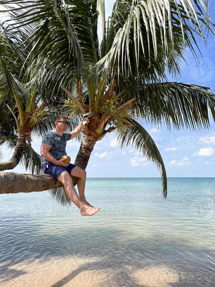 Man in sunglasses sits on a slanted palm tree holding a coconut in his hands. In the background is the turquoise sea, blue sky and tropical landscape of Phu Quoc Island in Vietnam. photo