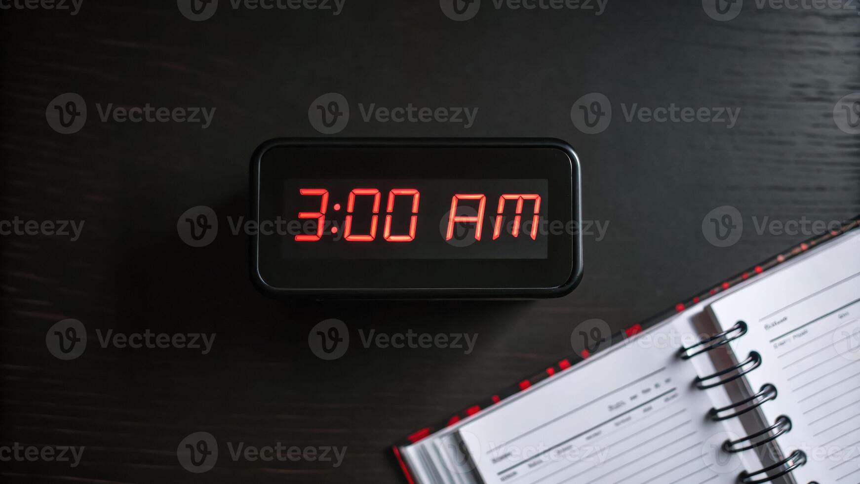 Digital clock 3AM red LED dark background planner open notebook time display early morning black surface spiral binding schedule organization photo
