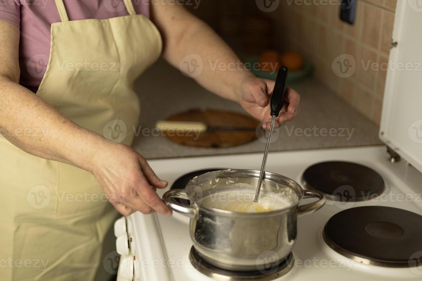 Senior woman's hands stirring sweet cream ghee with a metal spoon with holes in a saucepan on the stove photo
